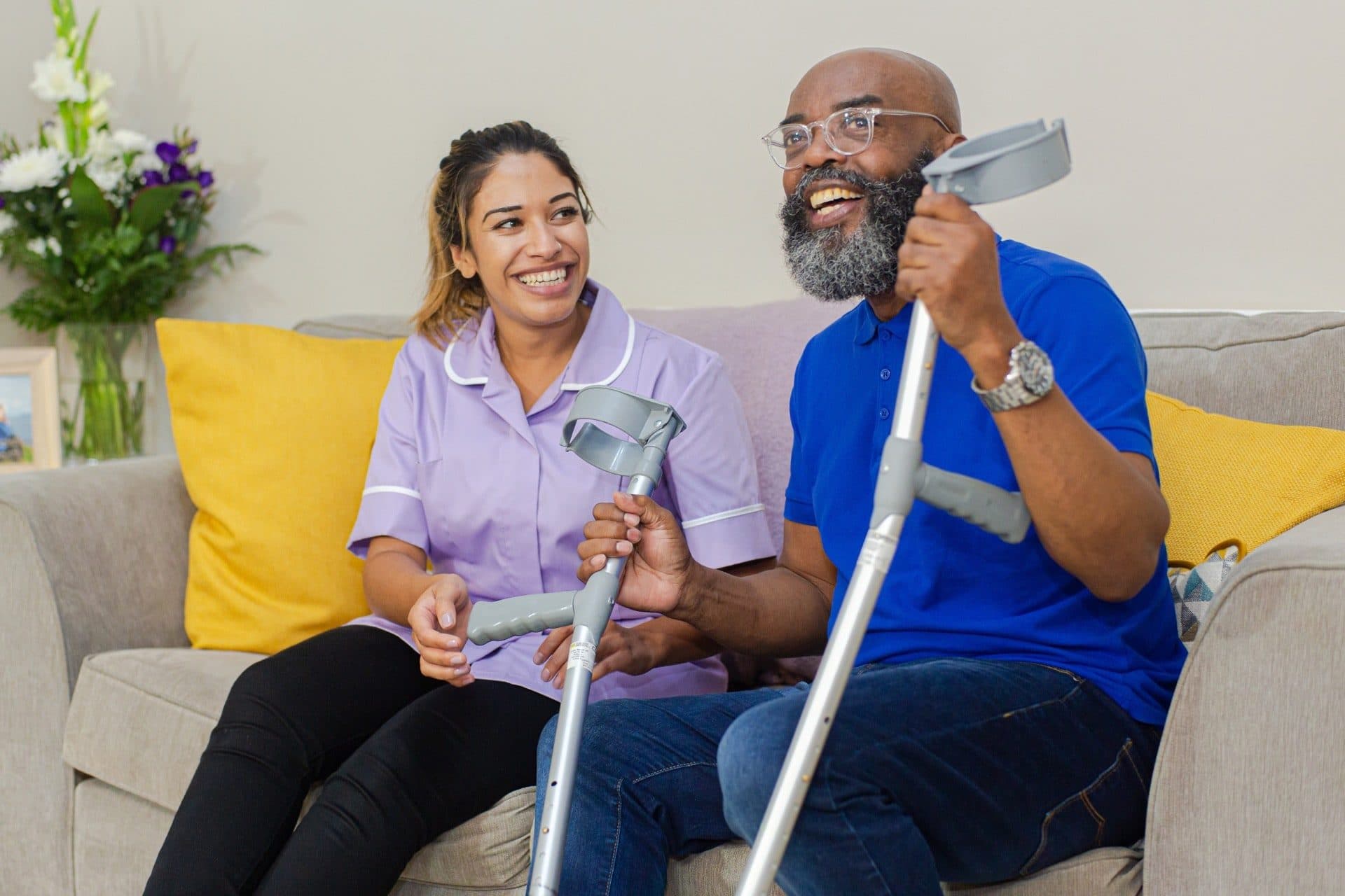 A carer laughing with a man using crutches on a sofa at home